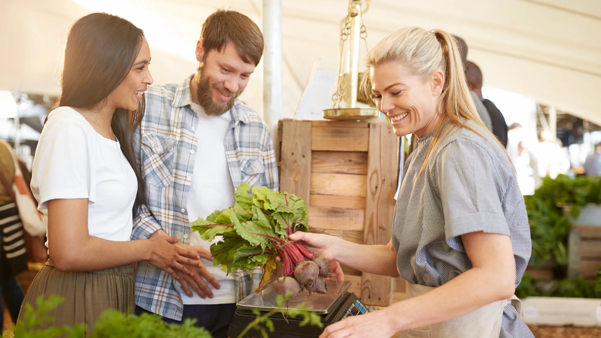 A grower at a market stall handing a bunch of freshly pulled beets across her scale to two shoppers